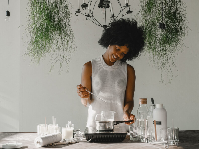A woman wearing white clothes stands at a table melting wax on a small burner