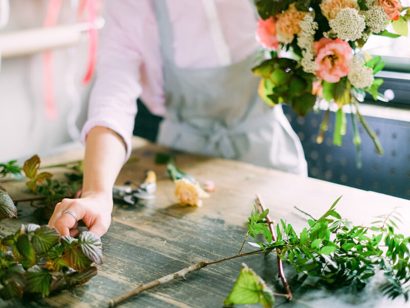 A person is standing at a table holding a bunch of white and orange flowers in one hand and is picking up green foliage with the other hand