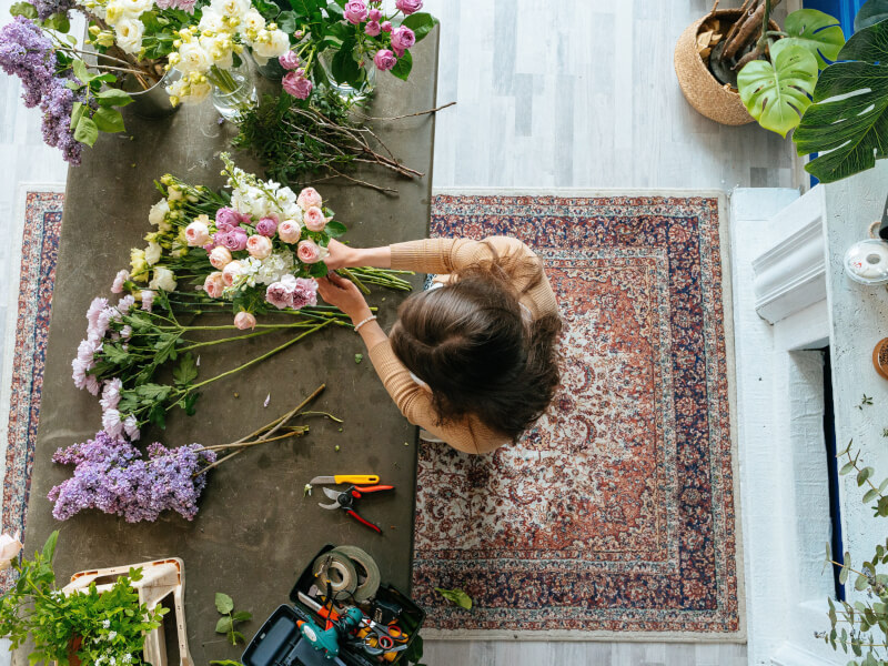 A top down shot of a person standing at a wooden table covered in a selection of flowers and greenery