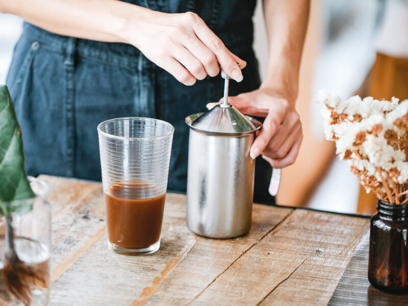 A person standing at a wooden bench making a cold brew coffee in a silver jug with a glass half filled with coffee next to it