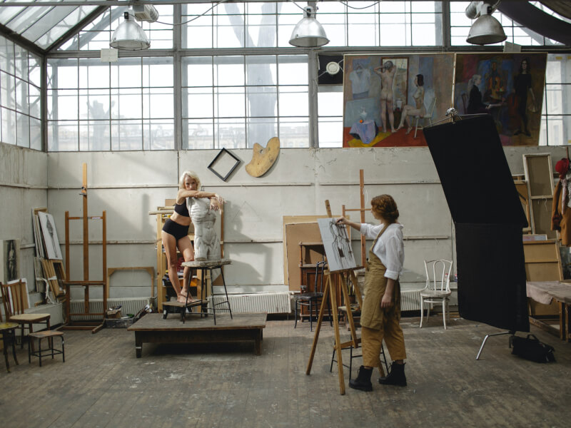 Person posing whilst a woman draws at a life drawing class.