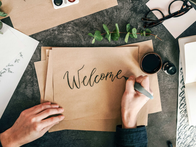 A person is writing 'welcome' in calligraphy on a brown envelope on a table. There is a eucalyptus branch and a cup on the table.