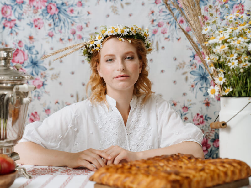 A female person wearing a small flower crown made from daisies, with a vase of flowers to her right side.