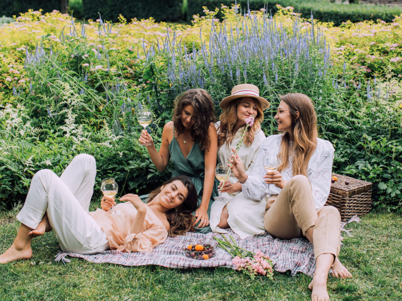Four girls sit on a picnic rug, laughing, with wine glasses in their hands
