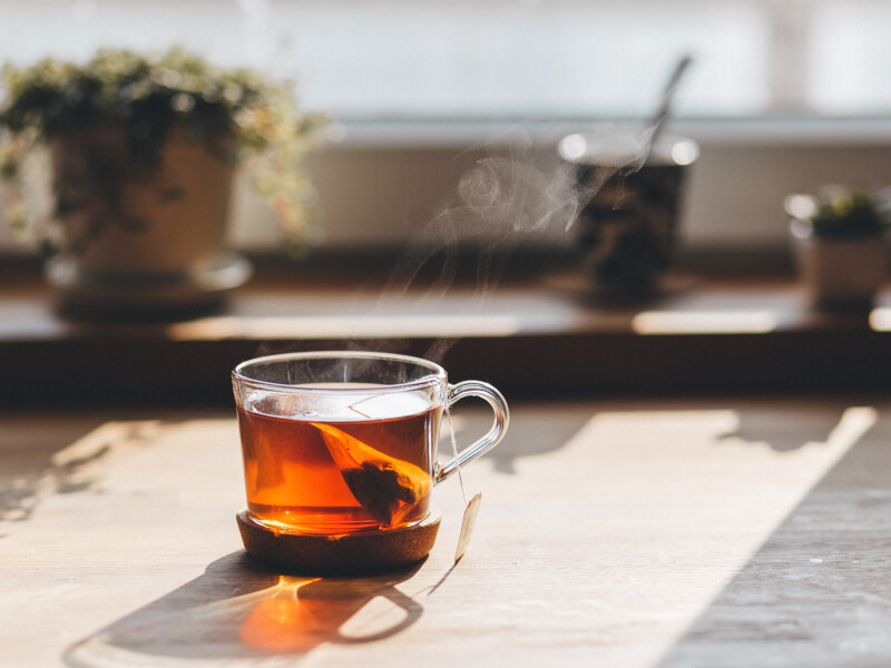 A close up shot of tea brewing in a glass cup. In the background, which is blurred, are two plants and a teacup.
