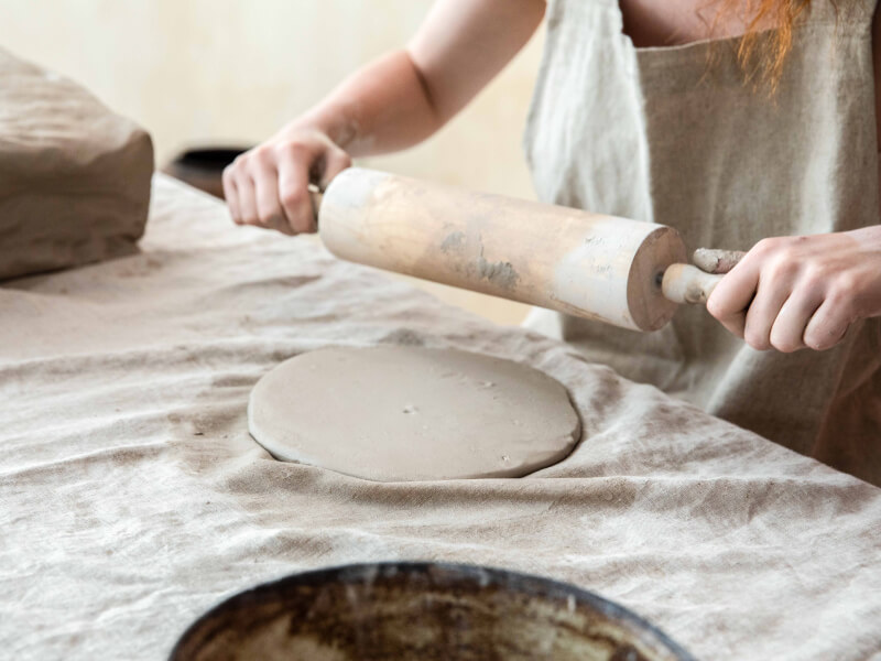 A flat piece of clay lies on a table, having just been rolled out by a person holding a rolling pin