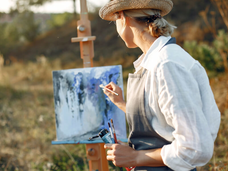 A woman is sitting at an easel outdoors, painting a landscape picture