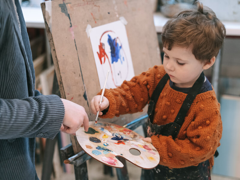 A teacher guiding a boy holding a paintbrush and palette.