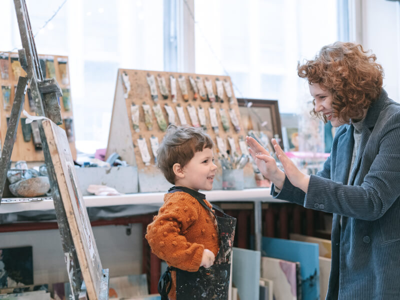 Woman teaching a toddler art next to easel.