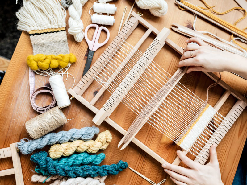 A person is weaving at a large wooden loom on a table. There is yarn, scissors and a tape measure on the table.
