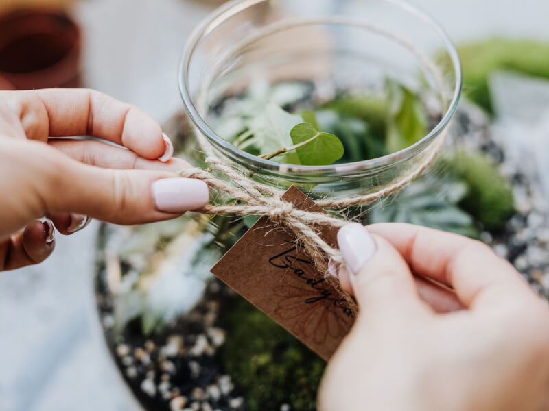 A person is hand tying a label around the neck of a glass terrarium