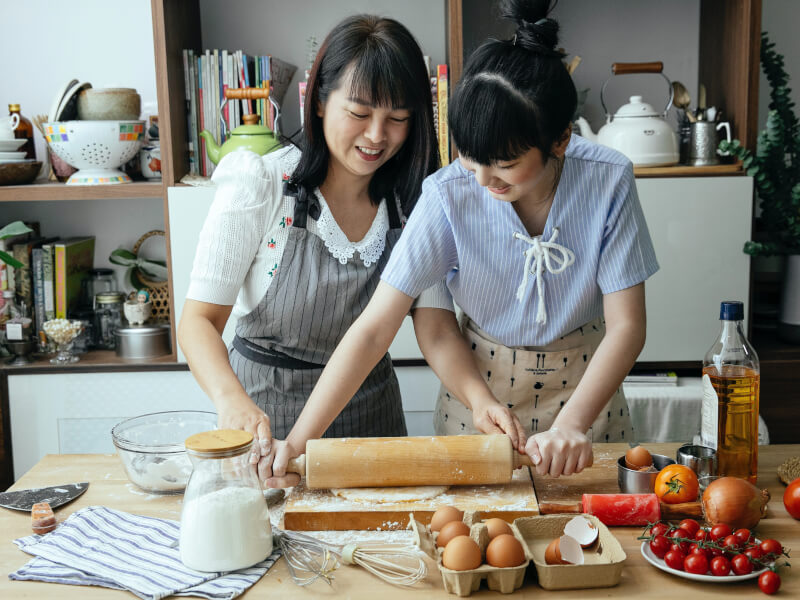 Two people using a rolling pin to roll dough as they take part in an online cooking class. A jar of flour, whisks, eggs and vegetables are on the table.