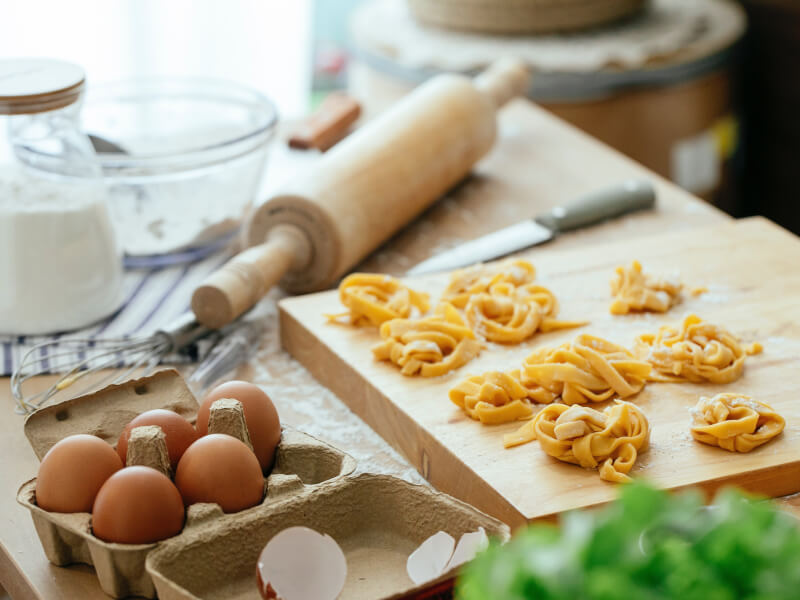Kitchen table covered in ingredients and tools for making pasta. There's a wooden chopping board with fresh tagliatelle on.