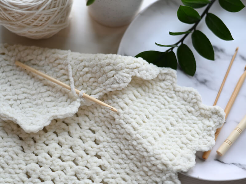 A piece of white crochet sits on a table with a crochet hook in it and some yarn nearby