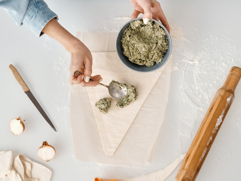 A person is spreading a green paste onto a flat triangle of pastry