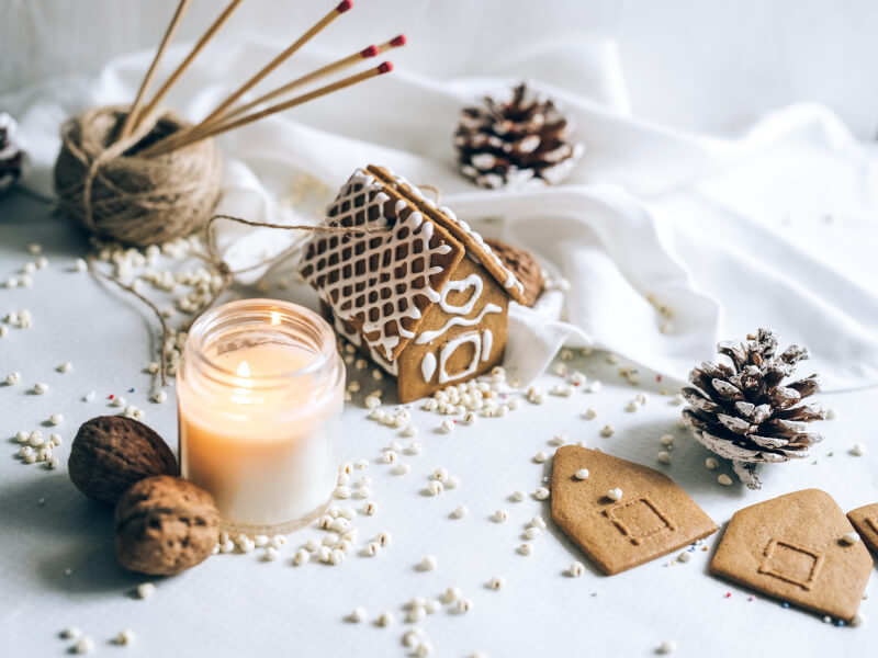 A lit candle and small gingerbread house sit on a white table with string and knitting needles in the background