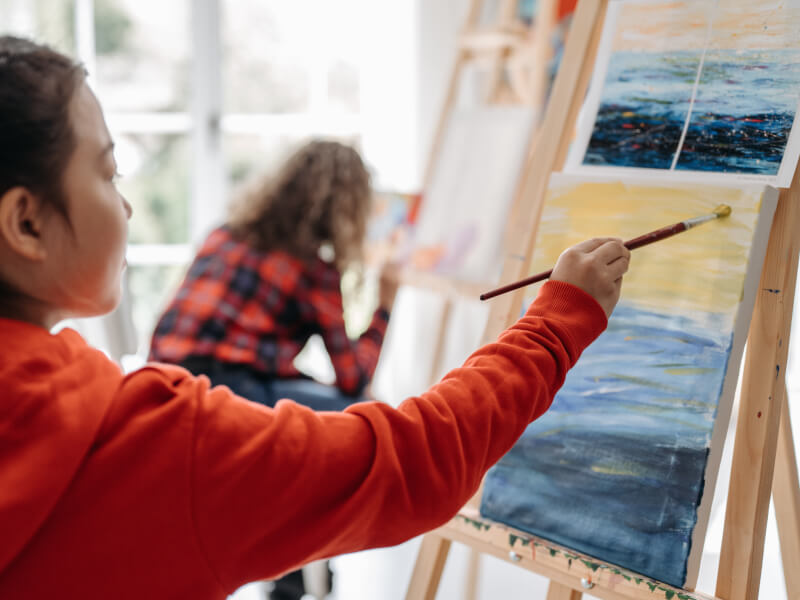 A woman in an orange jumper holds a brush as she paints on a canvas