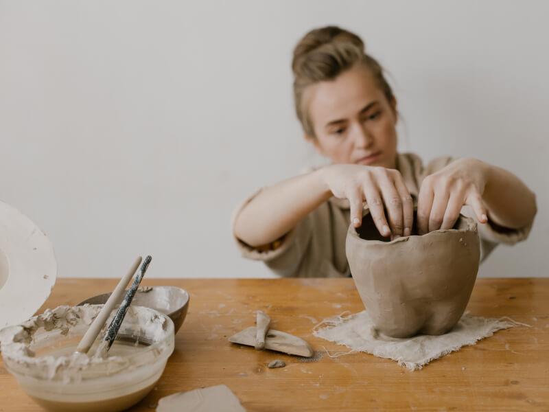 Woman sitting at a table shaping her pottery at home. Two bowls of water and pottery tools sit on the table.