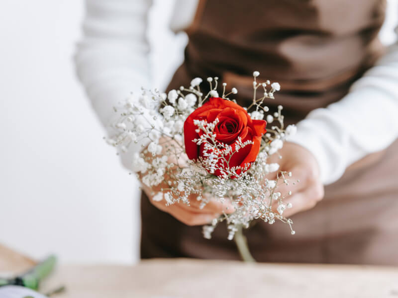 A person stands at a table holding a red rose and some white floral spray flowers