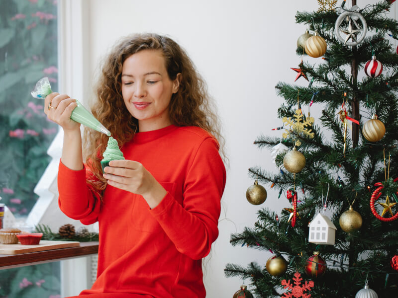 A woman in a red jumper is adding green frosting to a cupcake. There is a Christmas tree in the background