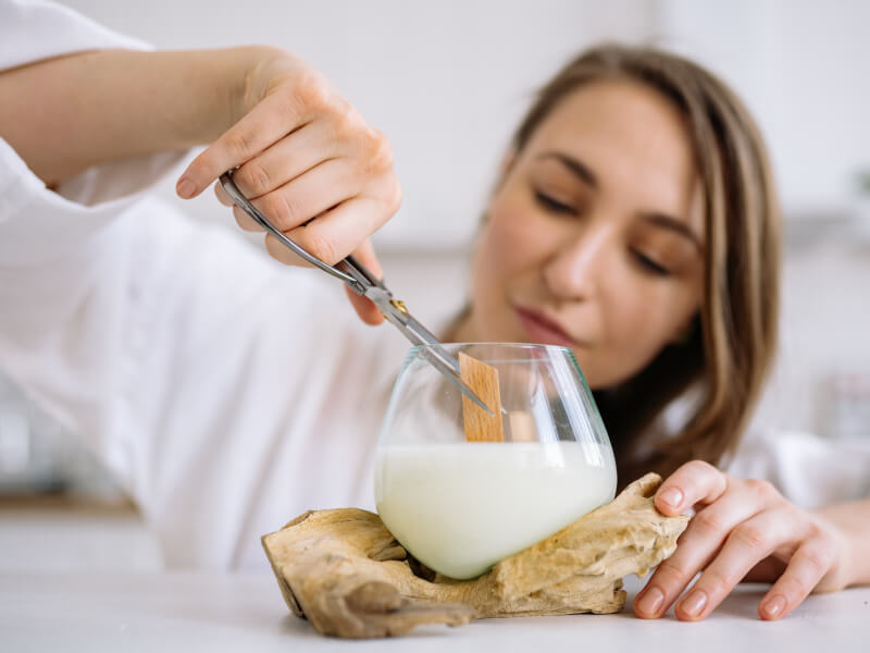 A woman is crouching behind a table, adding a wooden wick to a liquid candle in a glass pot