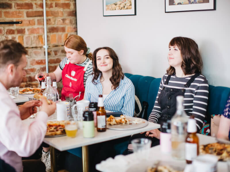 A woman smiles at the camera, sitting around a table with others at a pizza making class