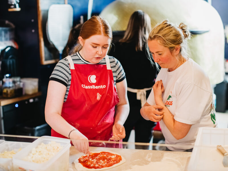 A cooking teacher showing a woman how to add sauce and toppings to a handmade pizza base