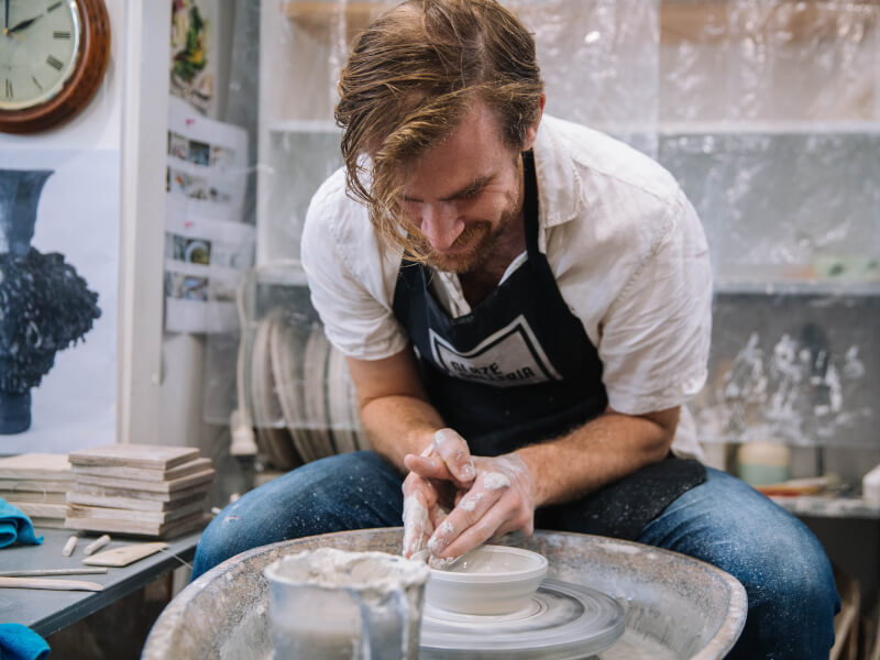 A man is sitting at a pottery wheel, making a small bowl shape out of clay
