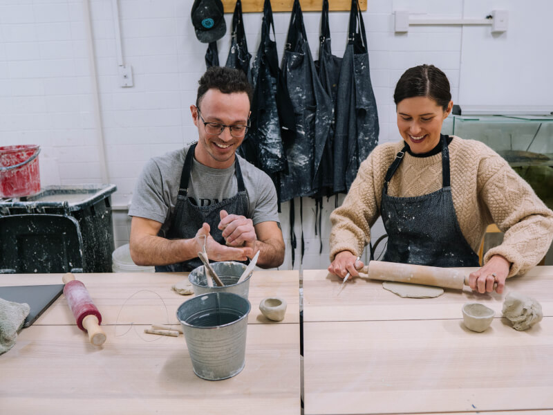 Two people sit at a wooden table, rolling out clay with a rolling pin on a wooden board