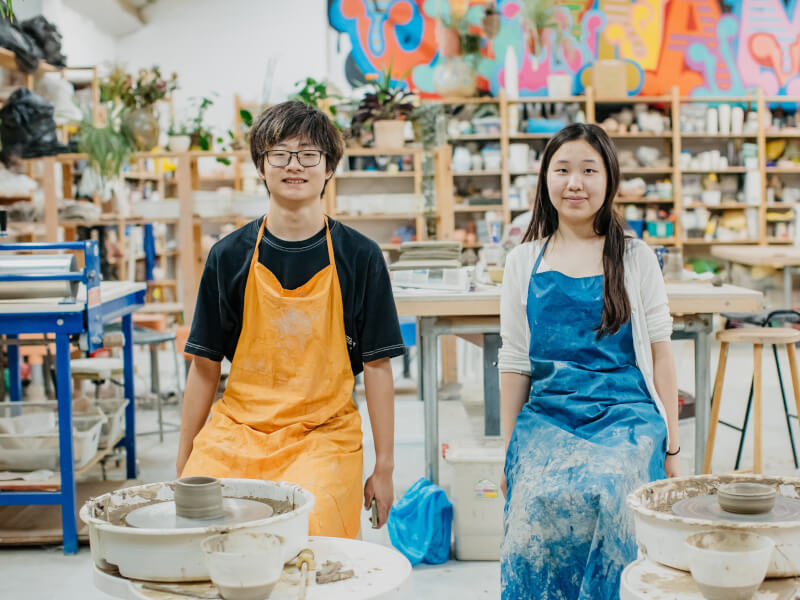 A man and woman sit at pottery wheels with freshly made pots, smiling