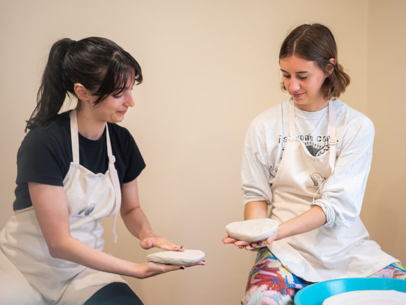 Two women are sitting at pottery wheels, showing each other the clay pots they've just made