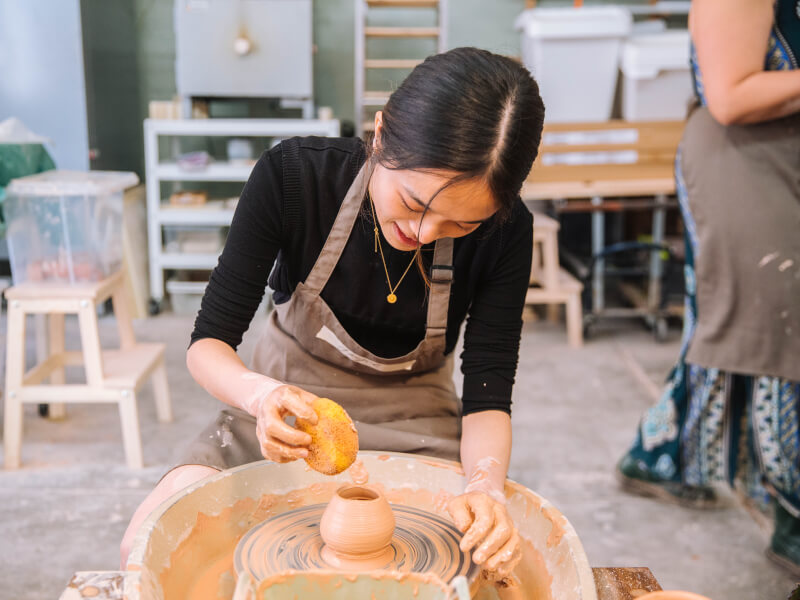 A woman is sitting at a pottery wheel, making a clay pot
