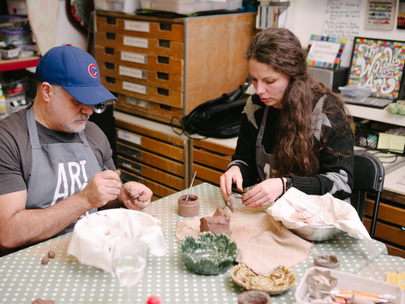 A man and woman sit at a table covered with green spotty tablecloth, hand building coil pots