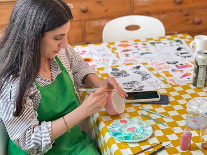 A woman is sitting at a table, painting a handmade ceramic cup pink