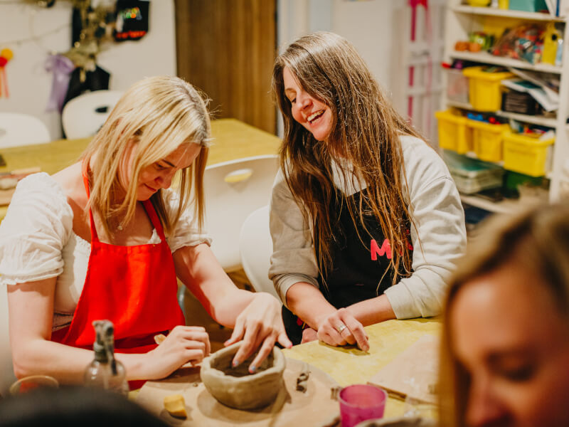 Two women smiling while shaping clay in a pottery class