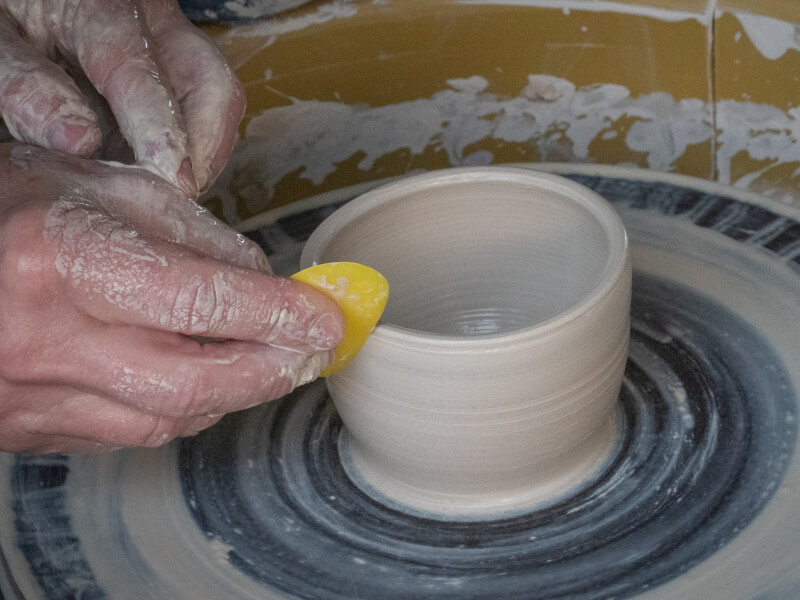 A person sits at a pottery wheel making a small bowl, using a tool to smooth the edges