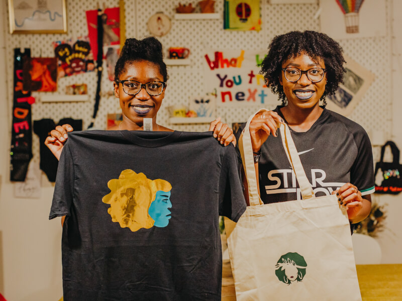 Two people standing up holding hand printed tote bag and tshirt