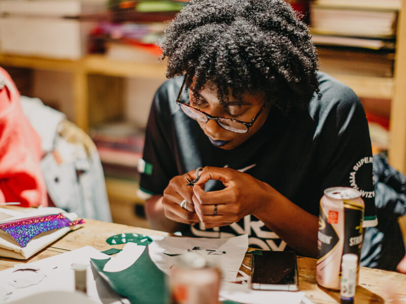 A woman is sitting at a table, cutting out a stencil to print onto a tote bag