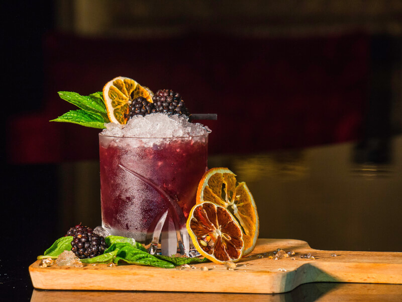 A cocktail in a glass tumbler with ice, dried oranges, mint and blackberries. Beneath is a coktail prep board.