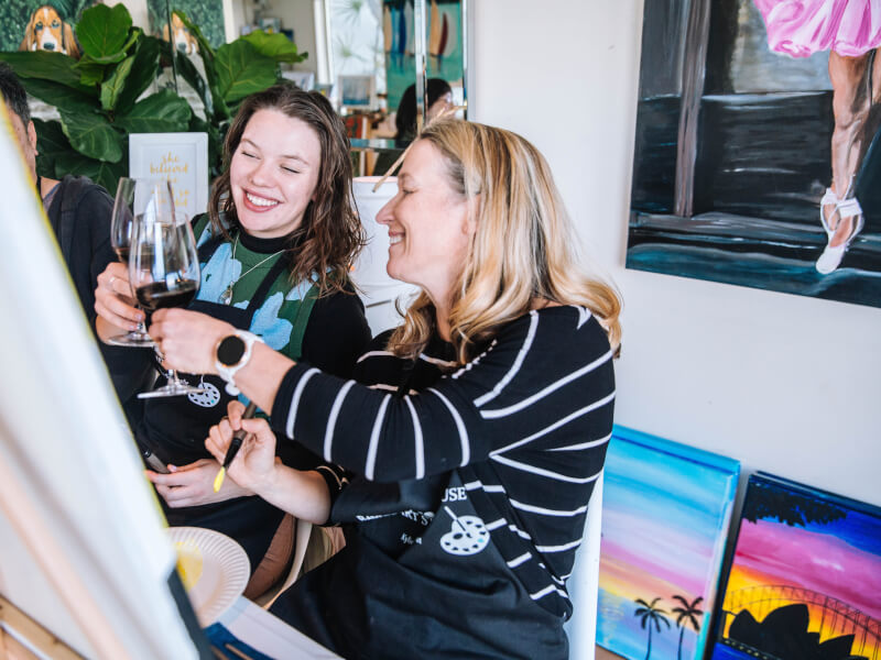 Two women are sitting at easels and clinking glasses at a painting class