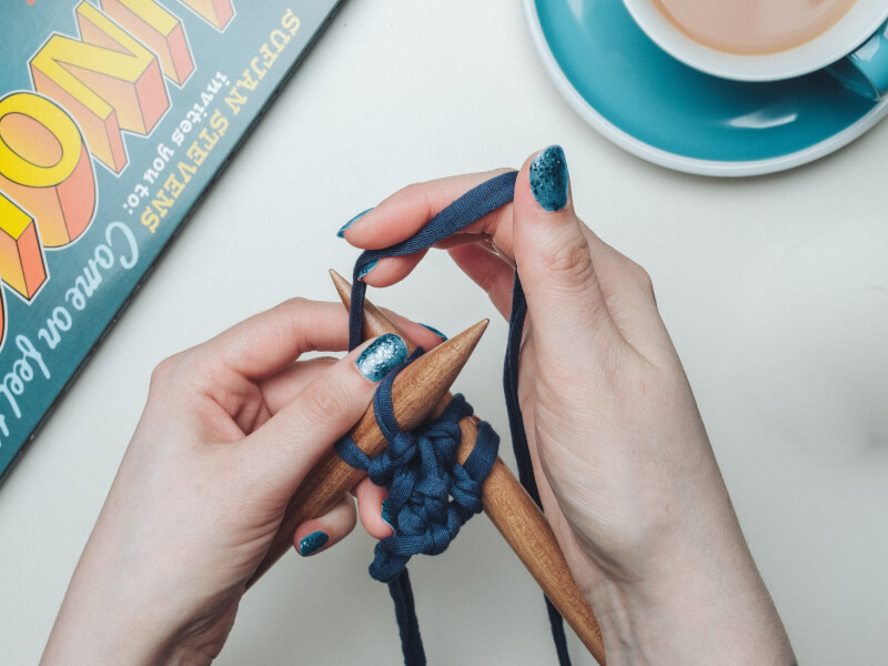 A person with blue nail polish is knitting with wooden needles in navy blue yarn