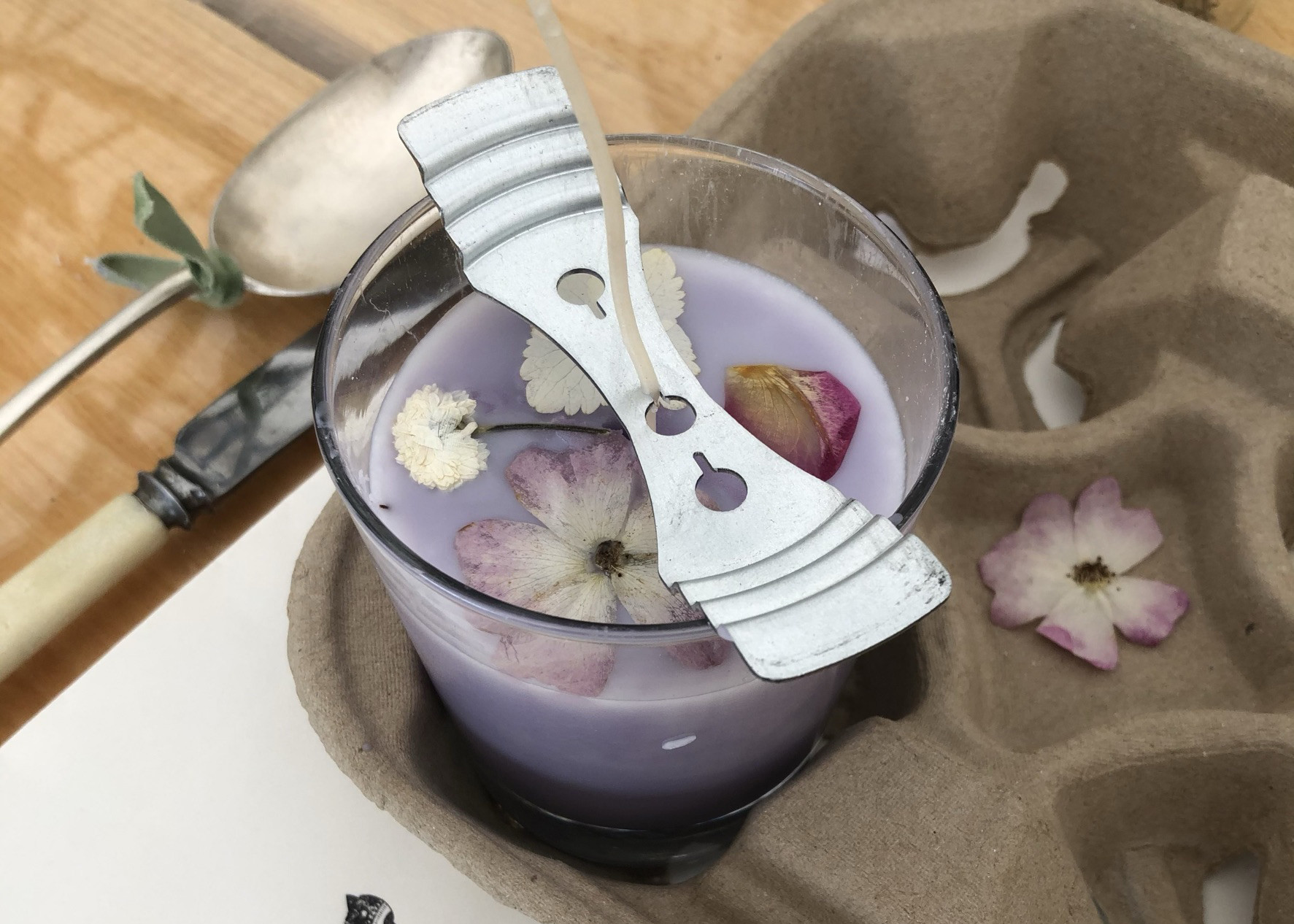 A handmade purple candle with flower petals on top is setting in a glass jar with a wick holder on top