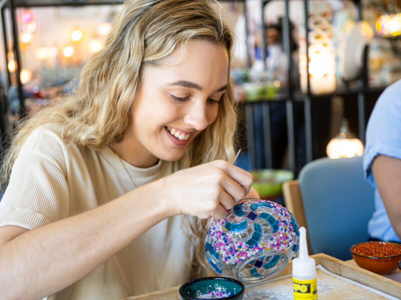 A woman is decorating a vase with mosaic tiles in blue, white and pink