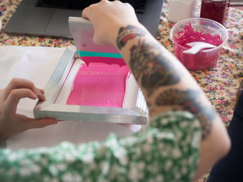 A close up of a person adding pink paint to a silk screen with a printing blade