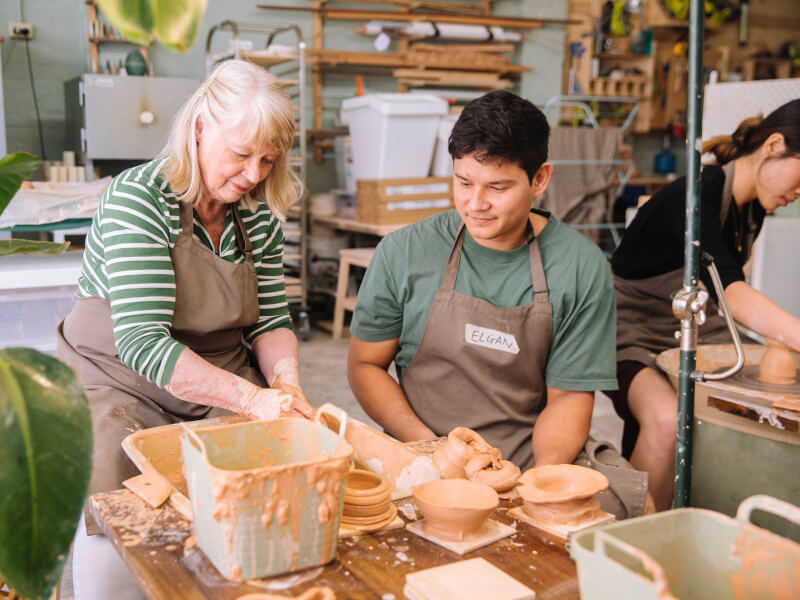 A lady is showing a man how to shape clay on a pottery wheel