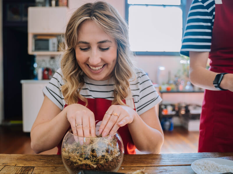 A woman is placing moss into a glass terrarium and smiling