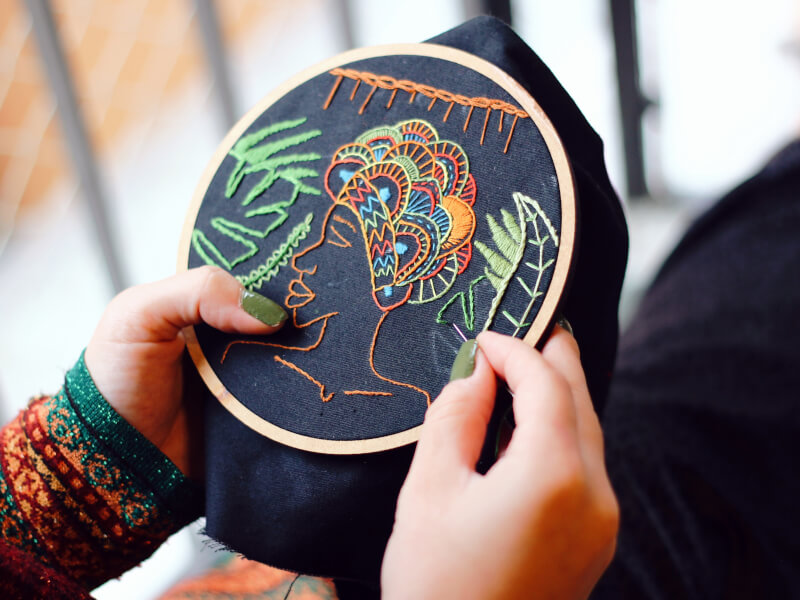 A person is sitting embroidering a colourful design on an embroidery hoop filled with fabric