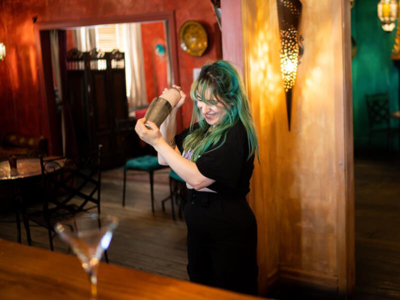 A smiling woman shakes a cocktail at shoulder height in a bar setting