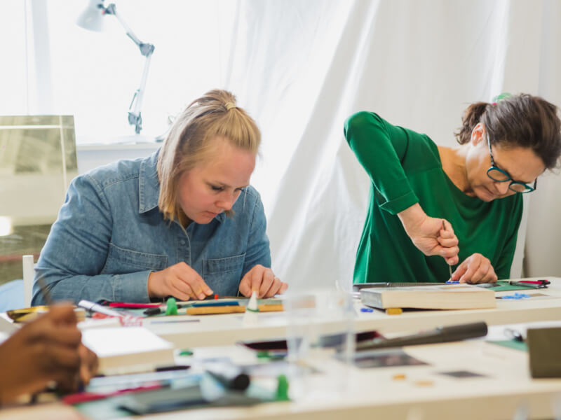 Two women are sitting at a desk using jewellery making tools.