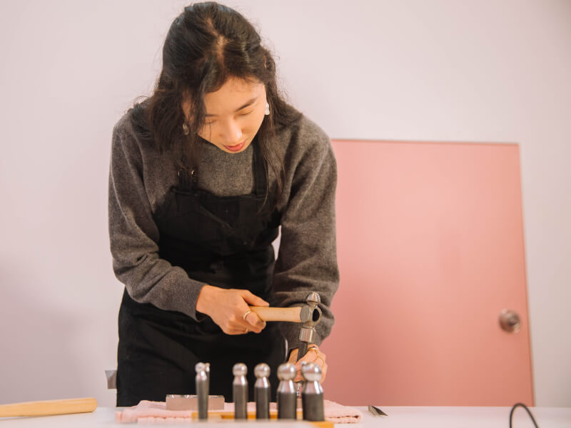 A woman is hammering a silversmithing project on a table, on top of a pink towel